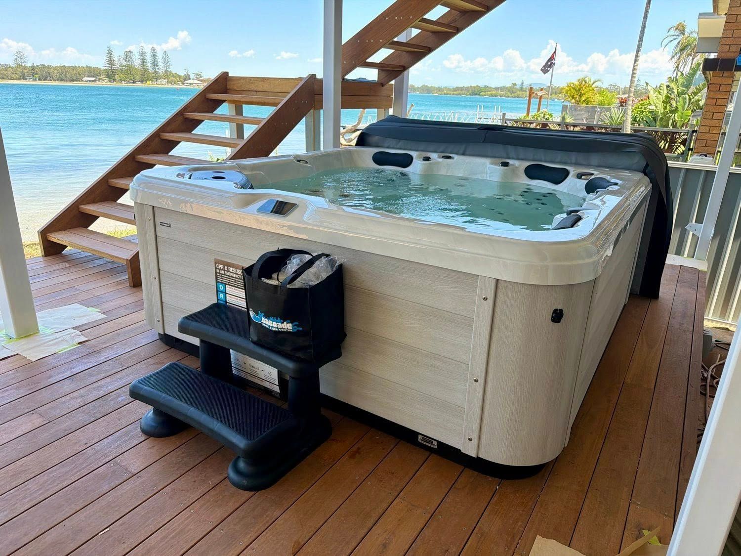 Hot tub on a wooden deck by the water, with stairs overhead and a small black step stool in front.— Cascade Pool & Spa Centre in Port Macquarie, NSW