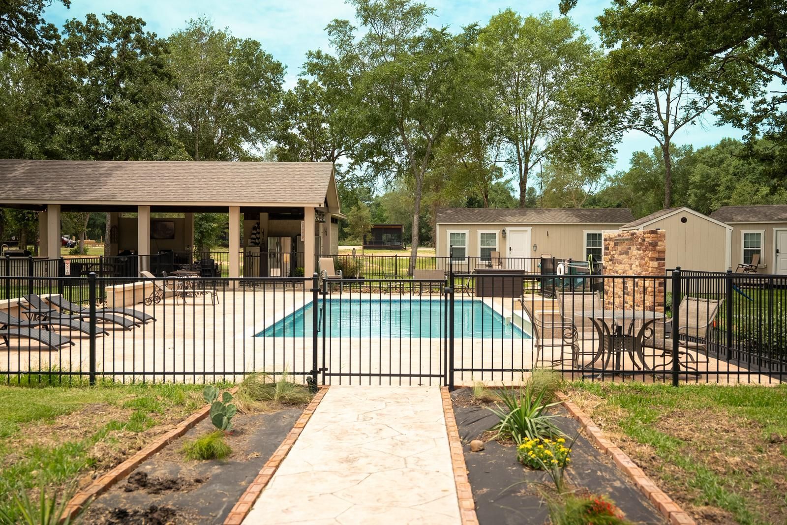 Backyard with a pool, patio, and small buildings. A black fence surrounds the pool and patio area, and there is a brick walkway.