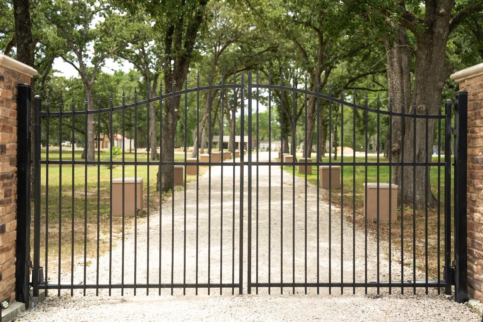 Black wrought iron gate between brick columns, leading to a gravel driveway lined with trees.