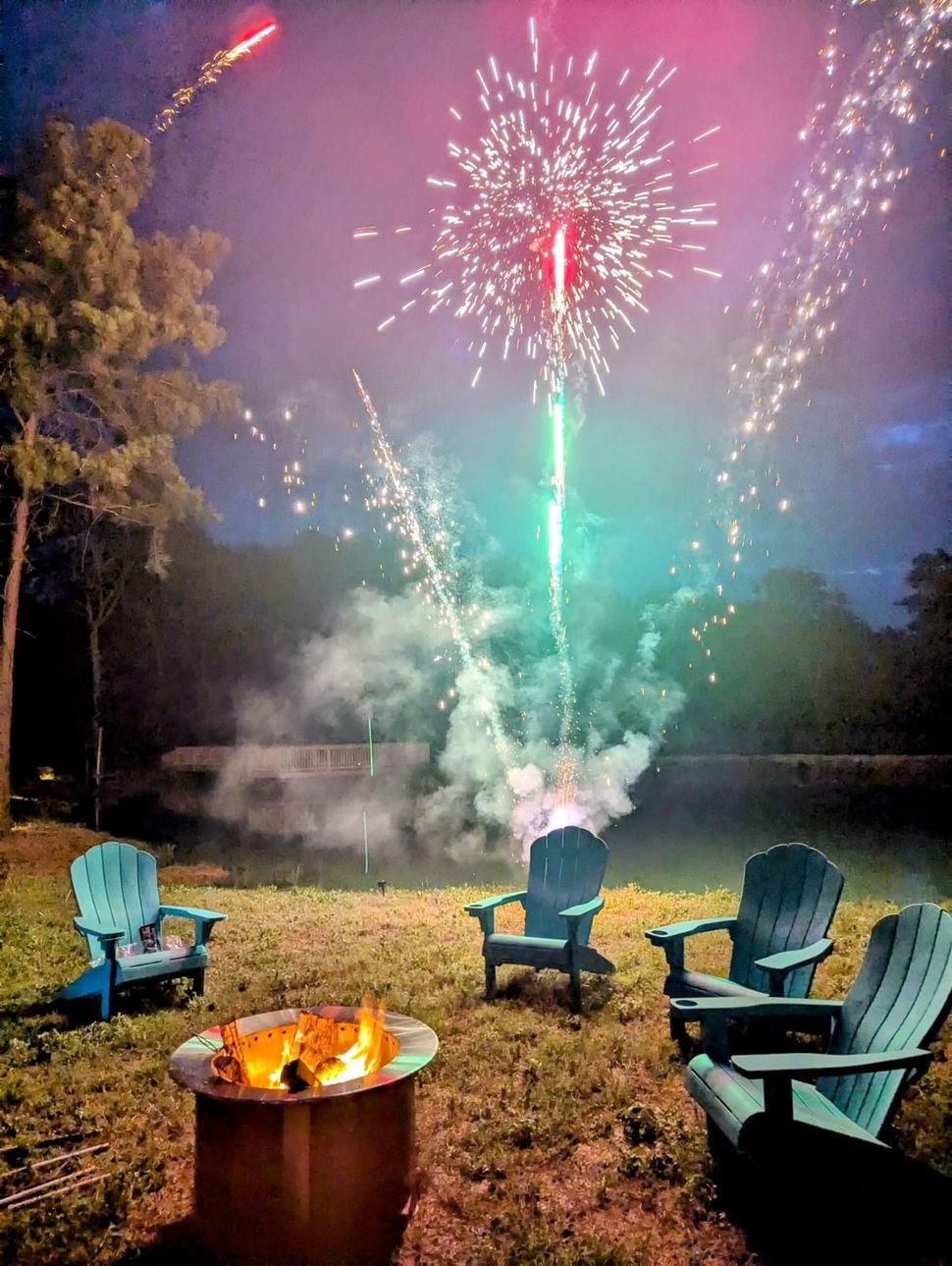 Fireworks over a fire pit with four Adirondack chairs in a backyard setting. Sparks and smoke fill the night sky.