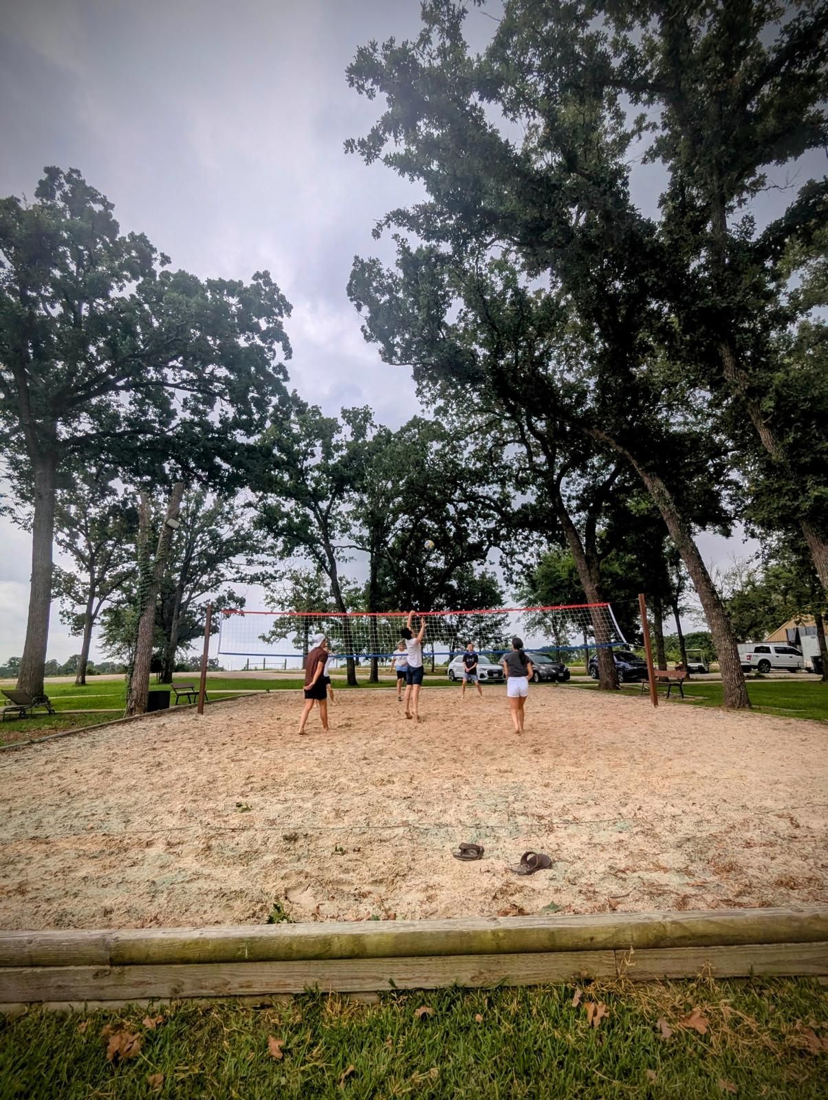 People playing volleyball on a sandy court, near trees and water under a cloudy sky.