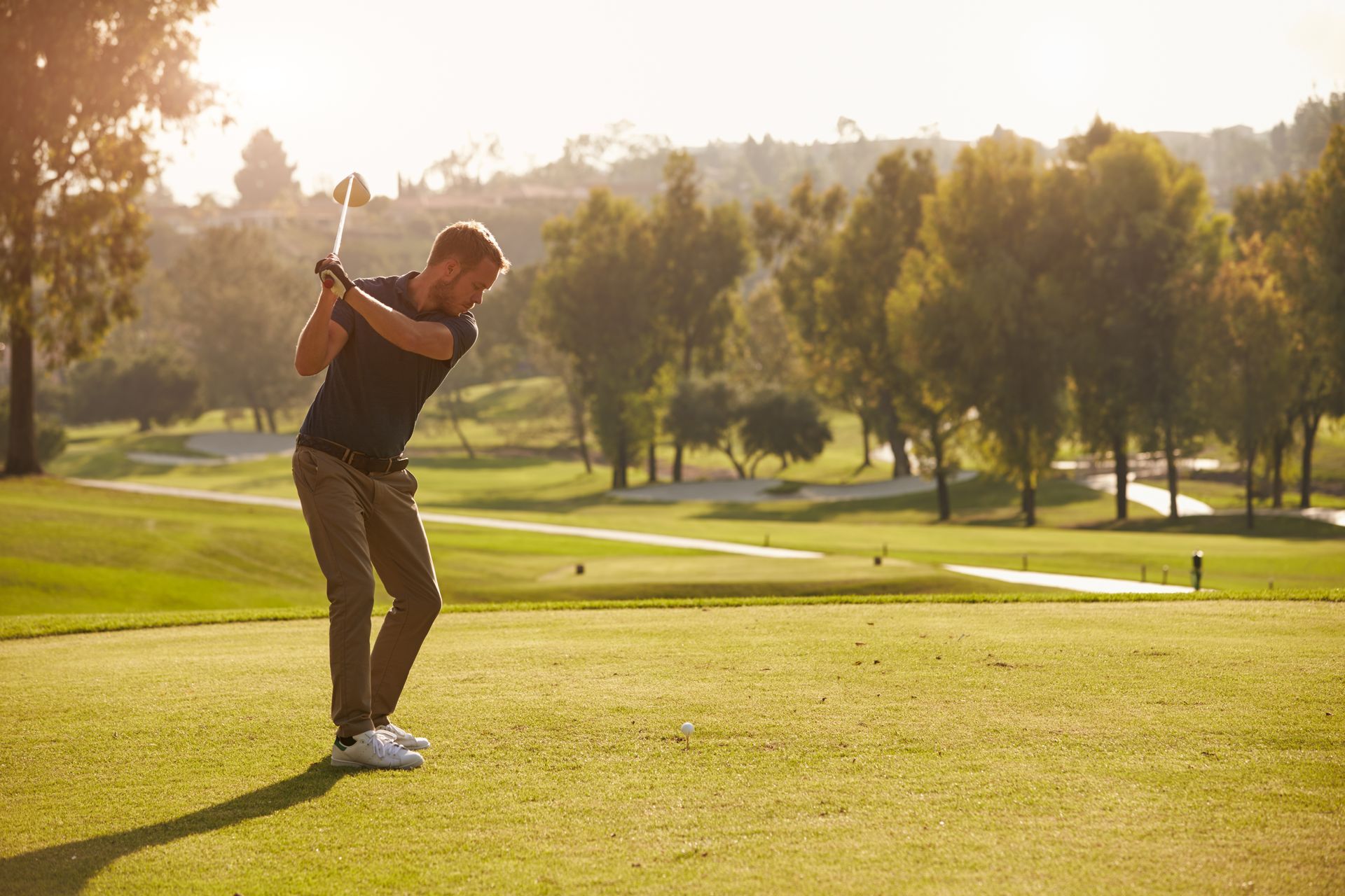 Man in dark shirt and khakis swinging a golf club on a green golf course in the sunshine.