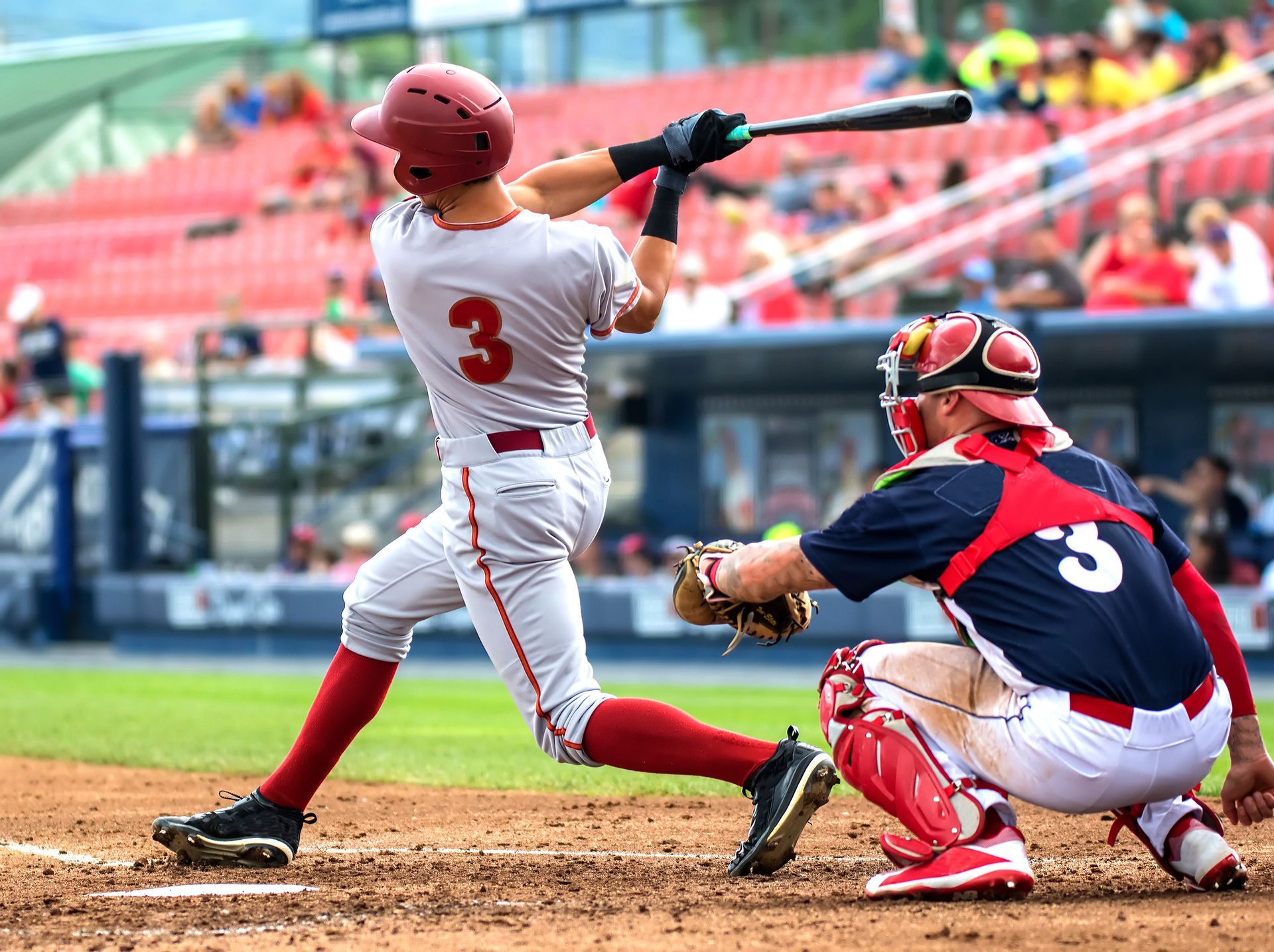 Baseball player in grey uniform swings bat; catcher in red and blue crouches. Stadium in background.