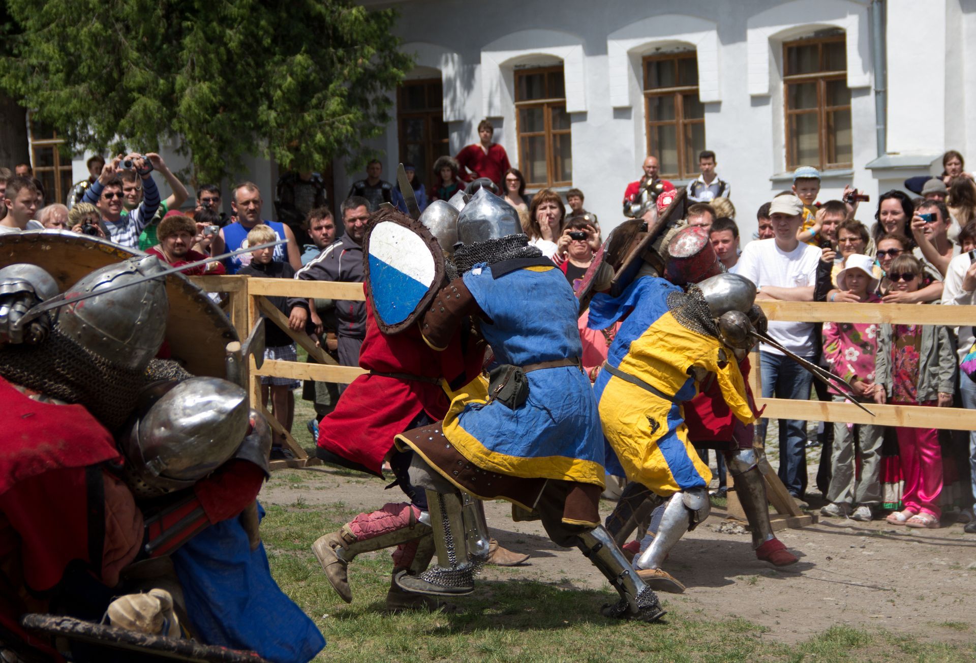 Medieval combat reenactment: Armored figures clash with swords and shields on a grassy field, surrounded by onlookers.