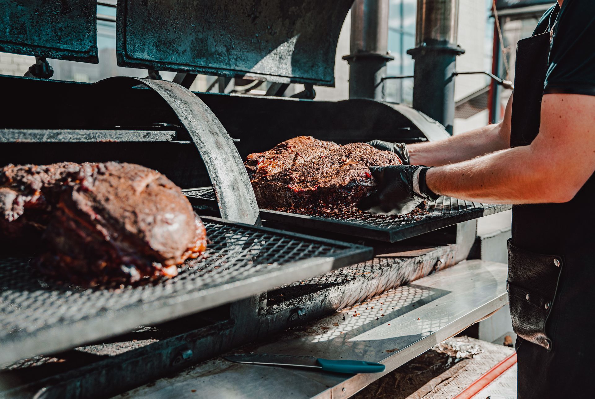 A person grilling large cuts of meat on a smoker. They are wearing black gloves and an apron in an outdoor setting.