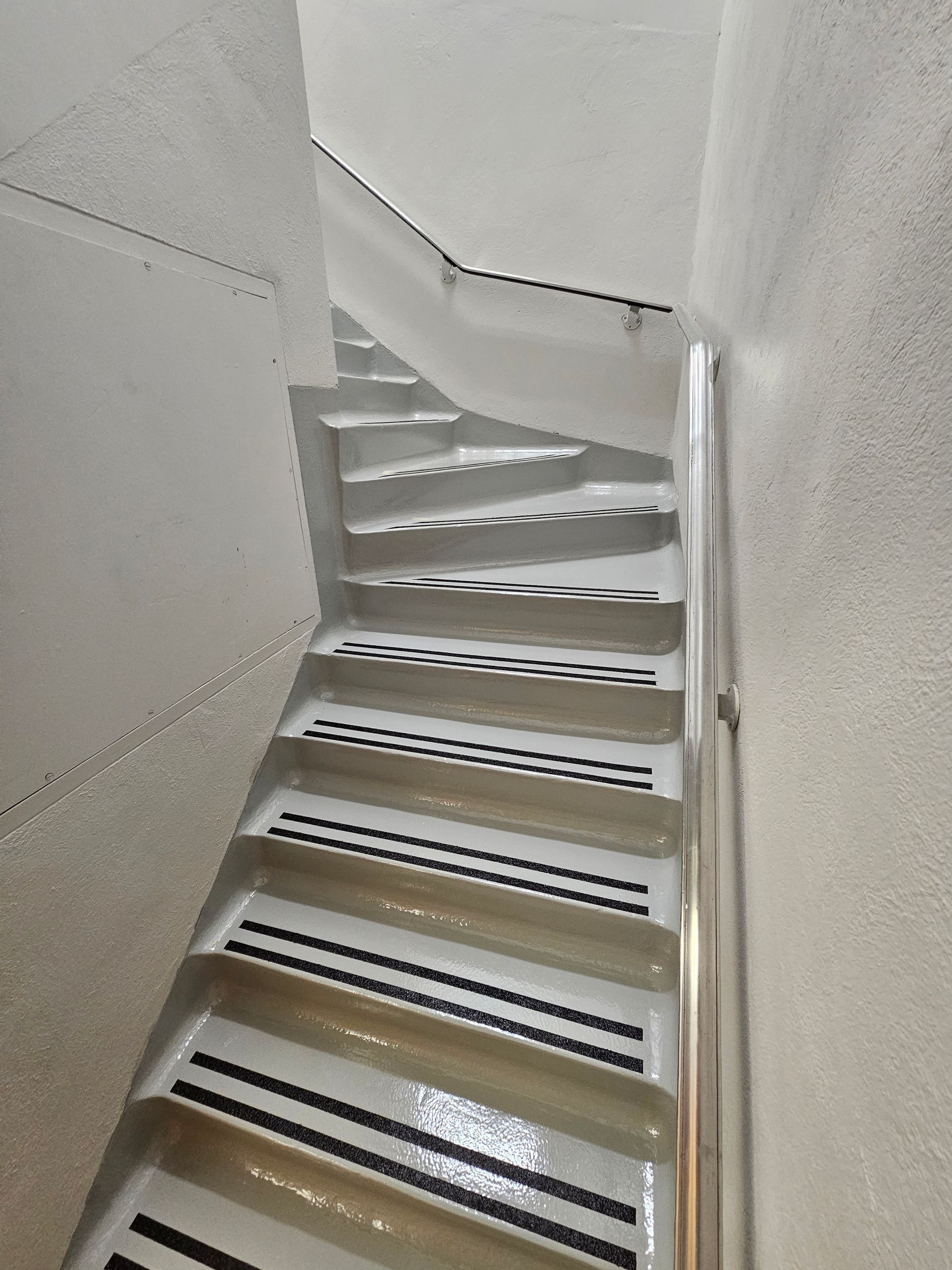 Escalier étroit et colimaçon blanc avec marches noires antidérapantes. Rampe argentée à droite.