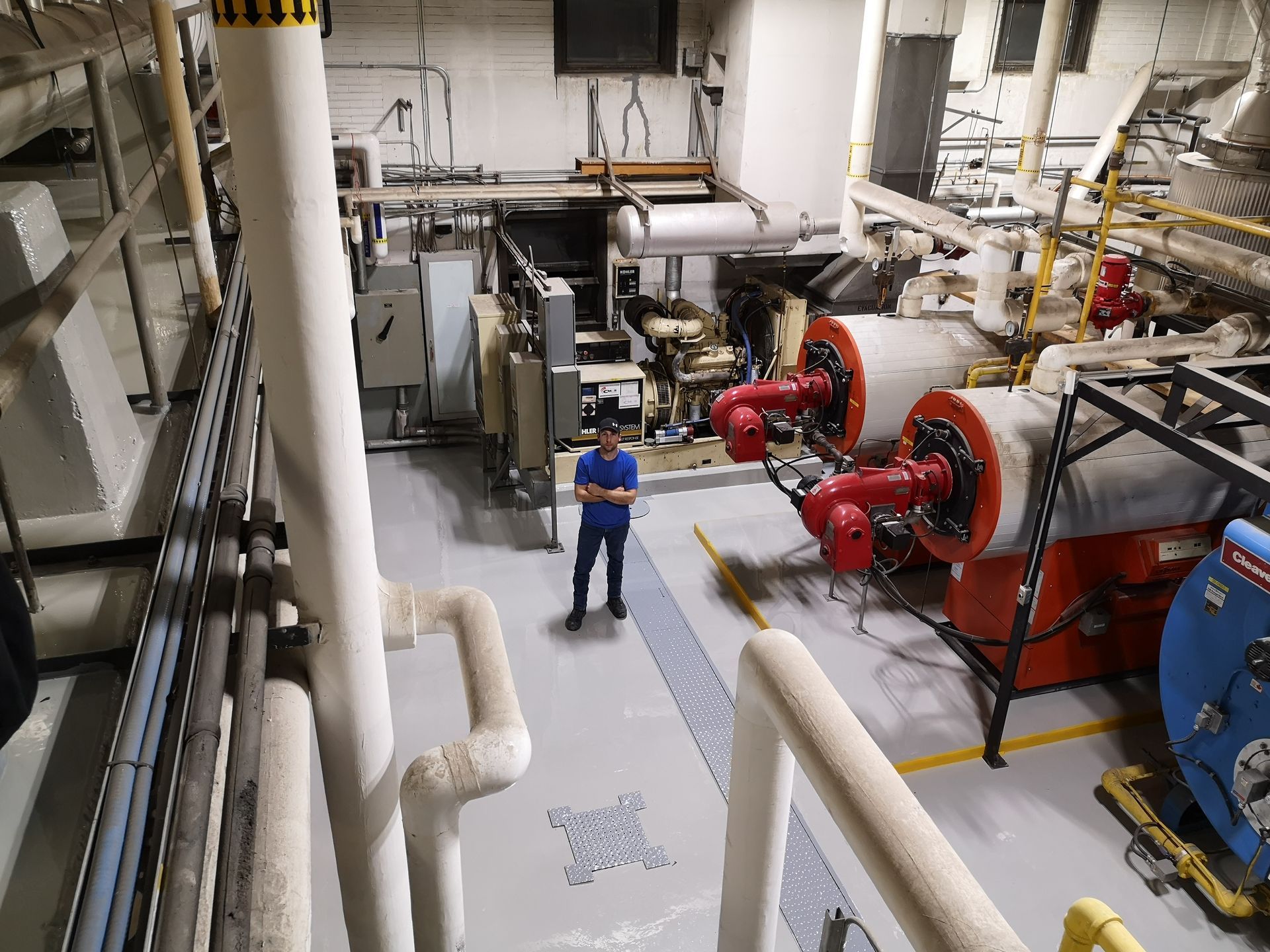 L'homme se tient dans une salle des machines avec de grands cylindres rouges et gris, des tuyaux blancs au-dessus.