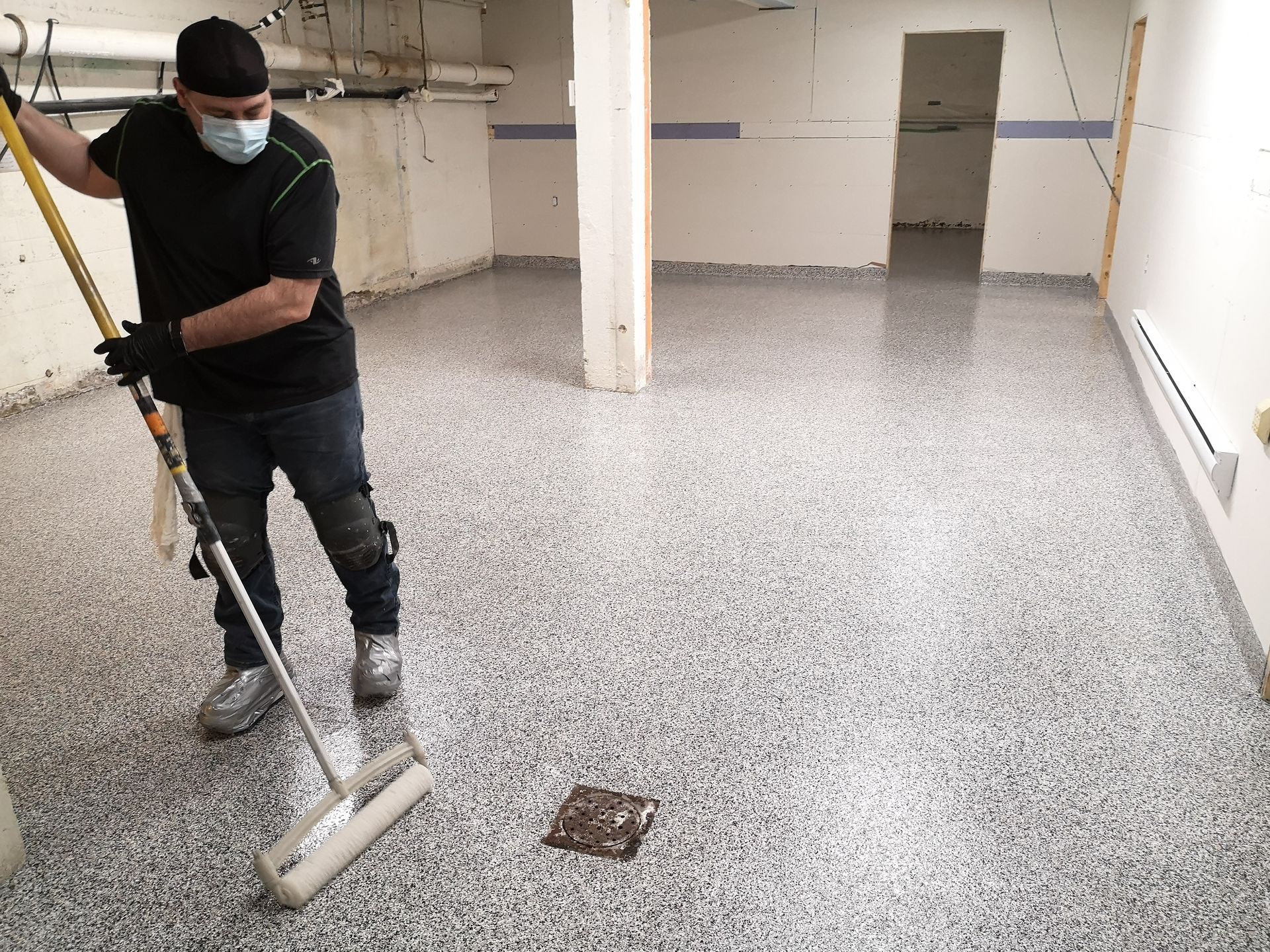Un homme masqué applique un revêtement de sol époxy dans une pièce. Sol moucheté gris et blanc, murs blancs.