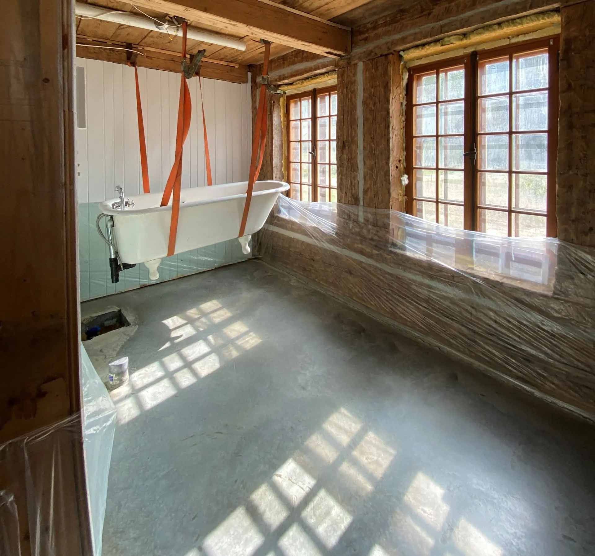 Une baignoire suspendue dans une pièce en travaux; fenêtres, sol en béton, sangles oranges retenant la baignoire.