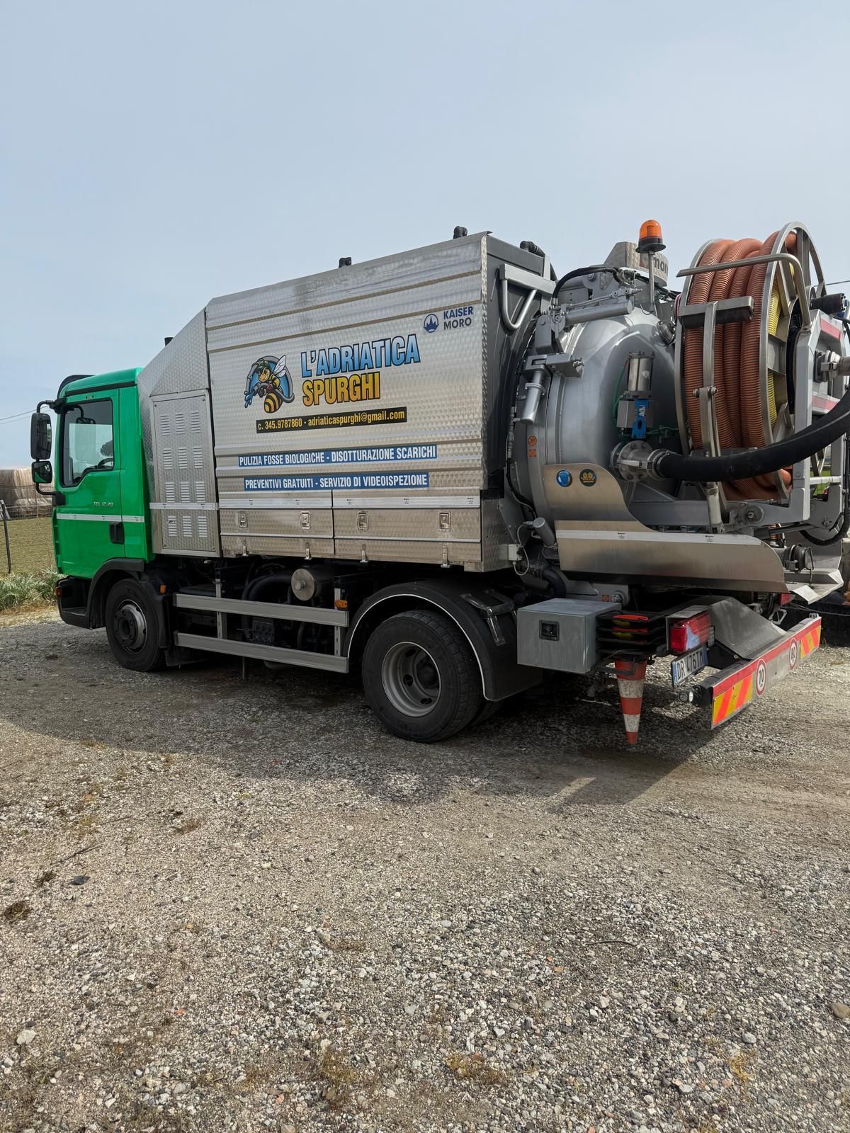 Un camion aspiratore per la pulizia delle fognature, di colore verde e argento, parcheggiato su una superficie ghiaiosa.