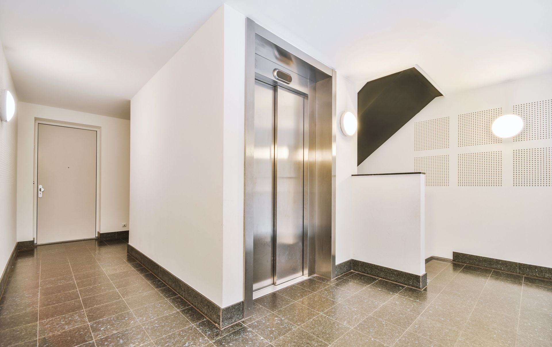 Una moderna hall interna con ascensore in acciaio inossidabile, pareti bianche e pavimento in piastrelle grigie screziate.