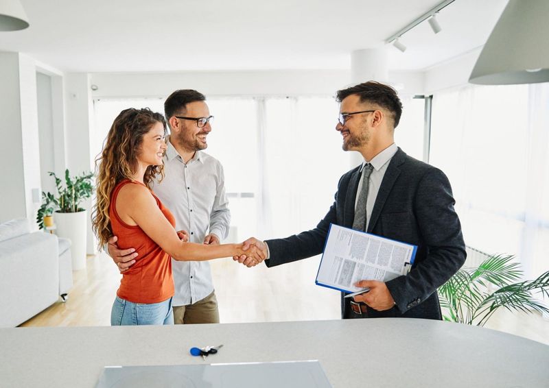 A man and woman are shaking hands with a real estate agent in a living room.