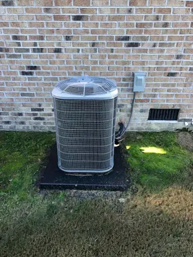 Air conditioning unit on a dark platform next to a brick wall and a small vent.