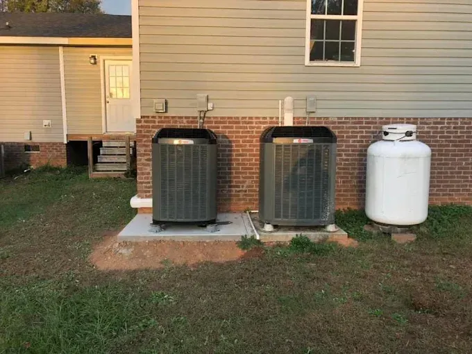 Two AC units and propane tank next to a house with brick and siding.