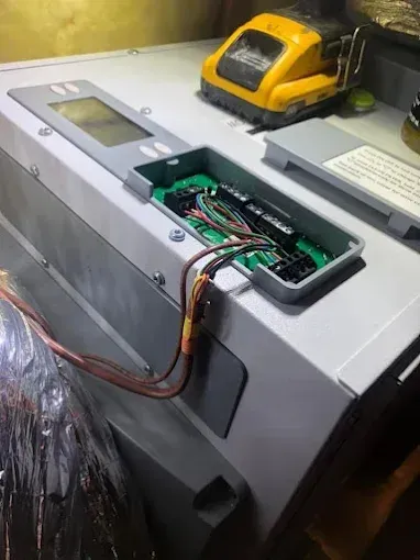 Open control panel on a gray HVAC unit, exposing circuit board and wiring. A yellow tool rests nearby.