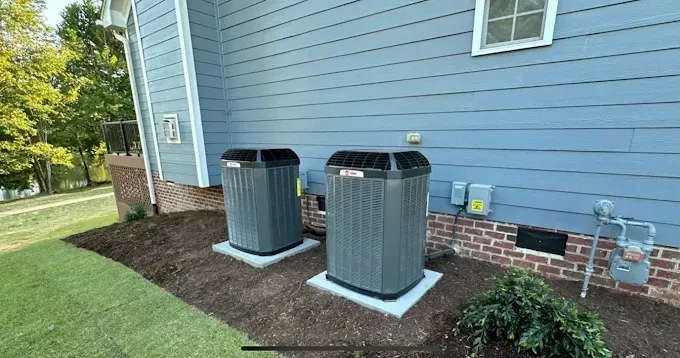 Two air conditioning units sit on concrete pads beside a blue house with grass and mulch.