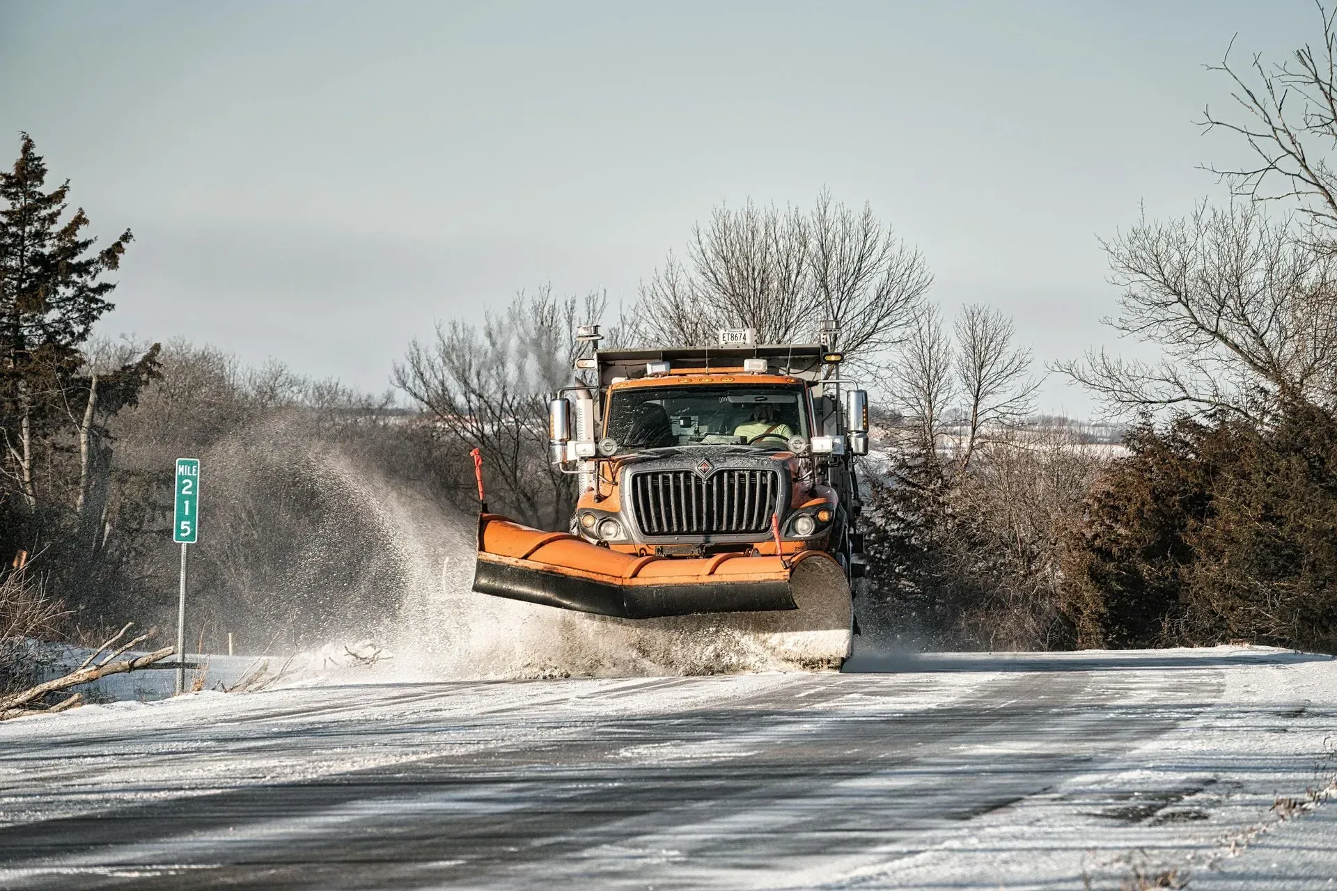 An orange snowplow clears snow from a rural road on a bright, sunny day.