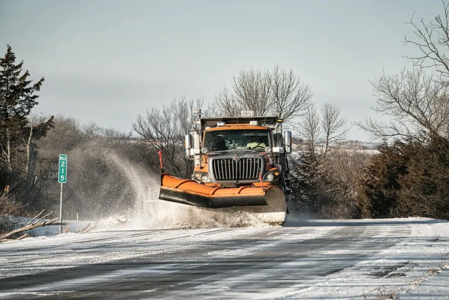 An orange snowplow clears snow from a rural road on a bright, sunny day.