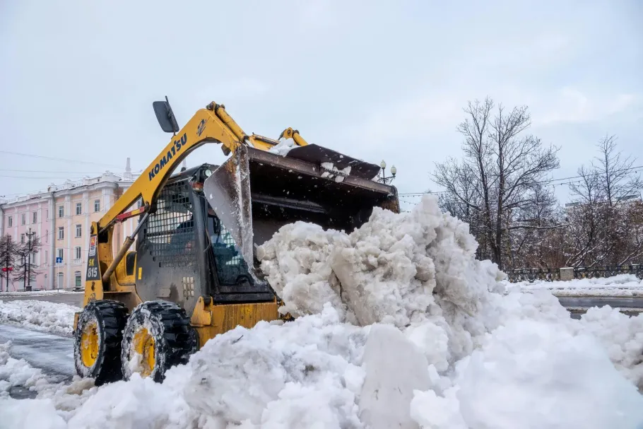 A yellow skid-steer loader clears a large pile of snow from a city street.