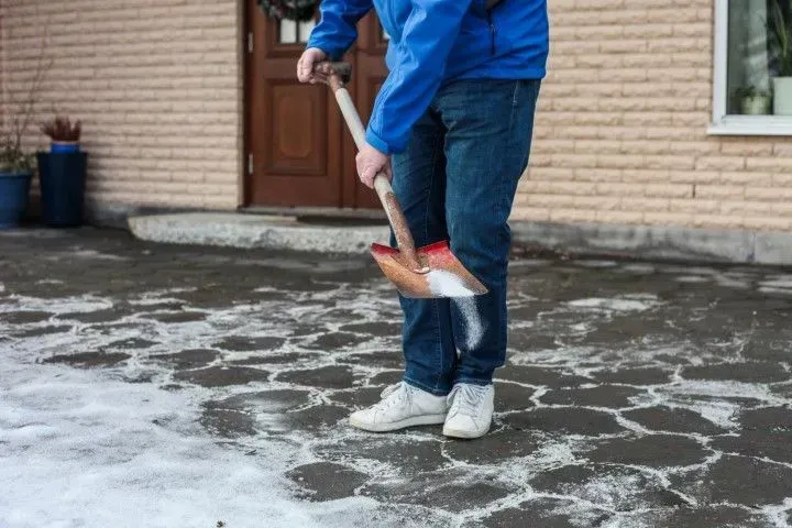 A person in a blue jacket uses a shovel to scatter salt on an icy paved area in front of a brick building.