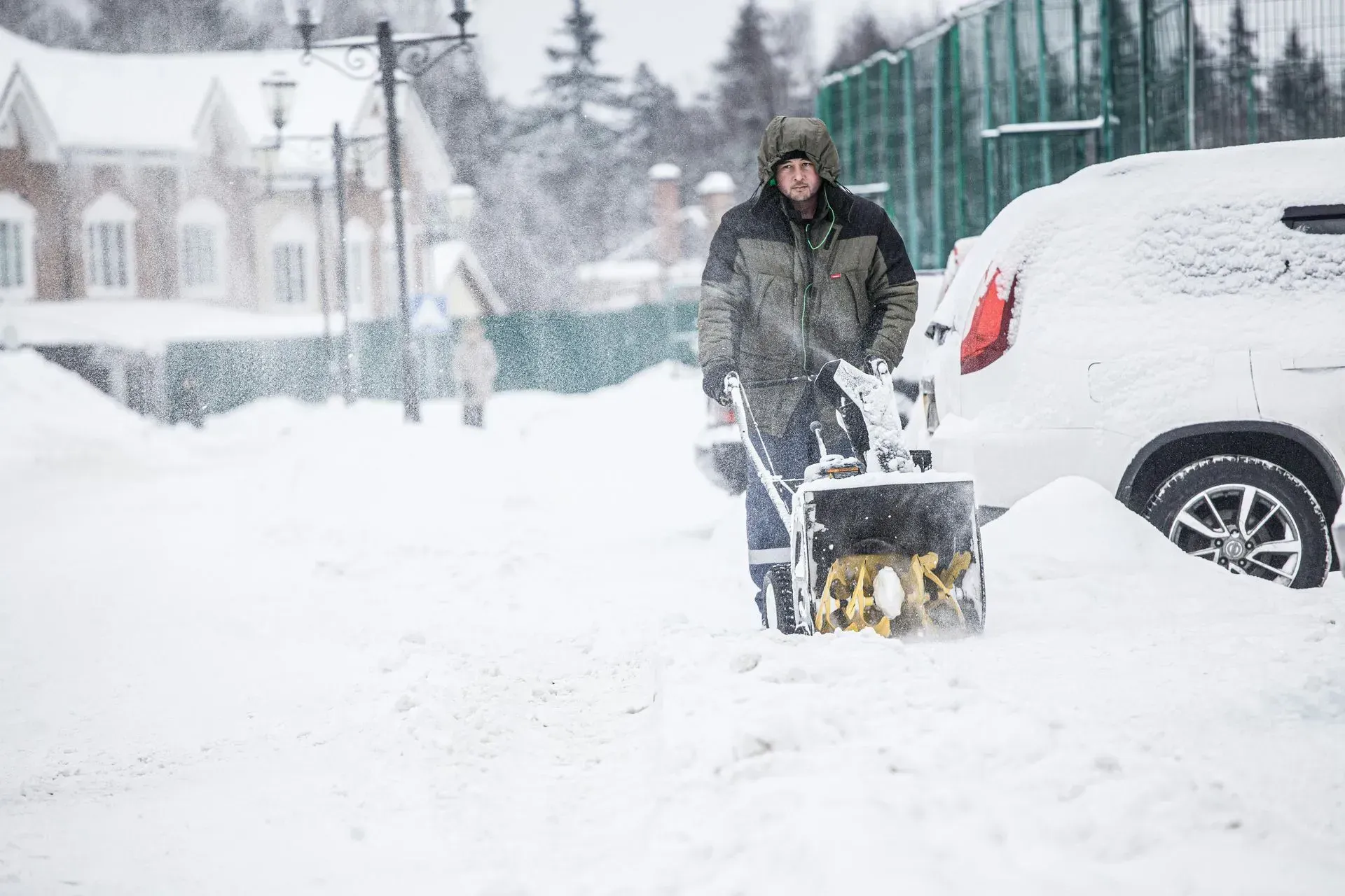 A person in a hooded winter jacket clears deep snow from a driveway with a yellow snow blower next to a parked car.