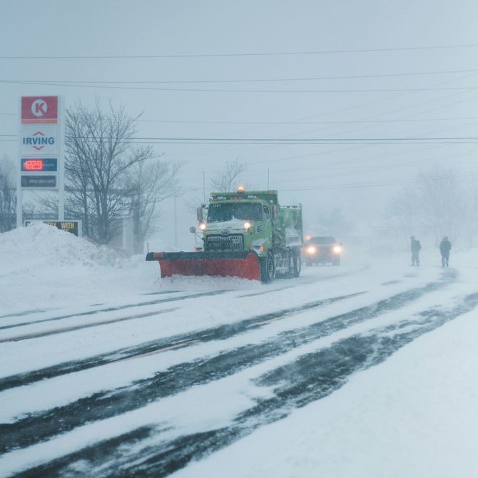 A snowplow clears a snow-covered road in front of a gas station during a storm as two people walk along the shoulder.