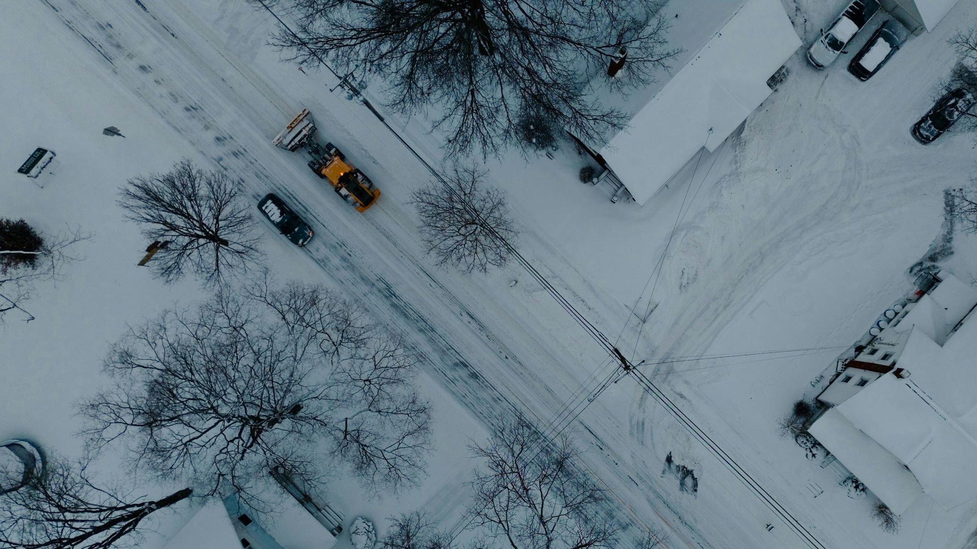 An aerial view of a bright yellow snowplow clearing a snow-covered street in a quiet residential neighborhood.