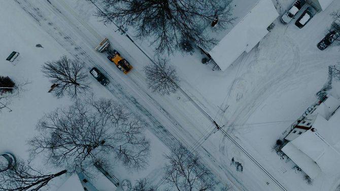 An aerial view of a bright yellow snowplow clearing a snow-covered street in a quiet residential neighborhood.