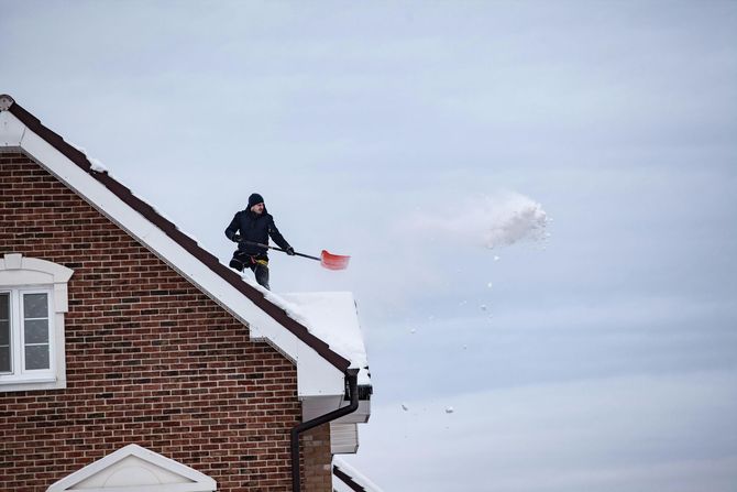A person on a brick house roof uses a red shovel to toss a large clump of snow into the air.