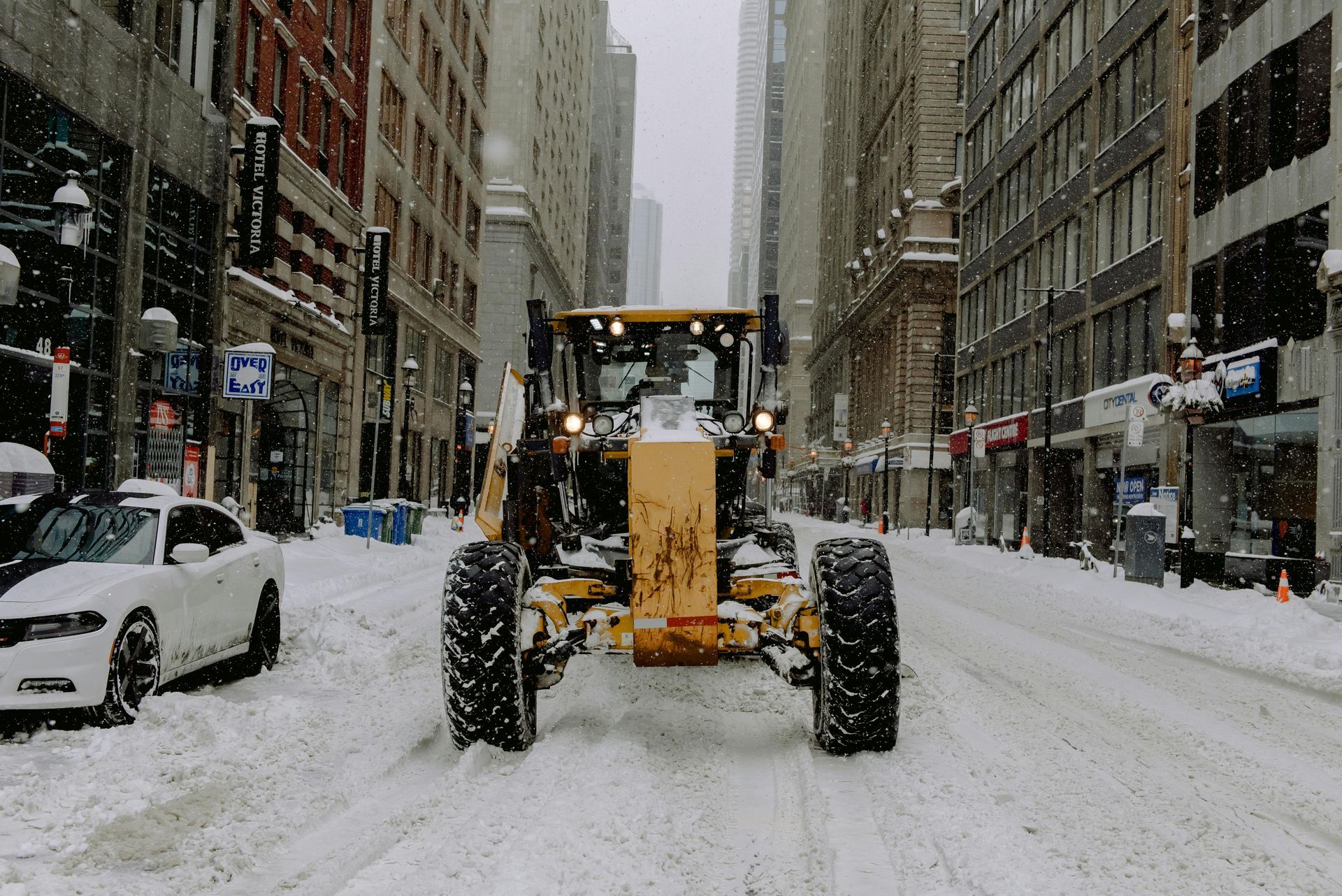 A yellow motor grader moves down a snowy city street between tall buildings, with a parked car on the left.