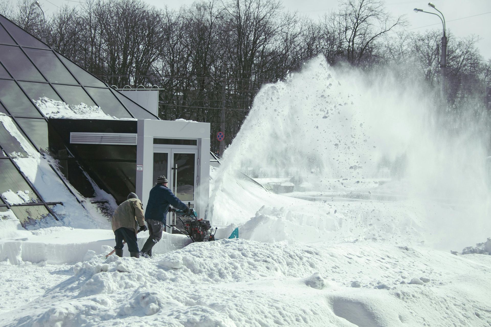 Two people use a snowblower to clear heavy snow outside a building with glass walls.