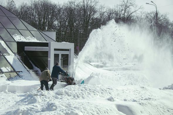 Two people use a snowblower to clear heavy snow outside a building with glass walls.