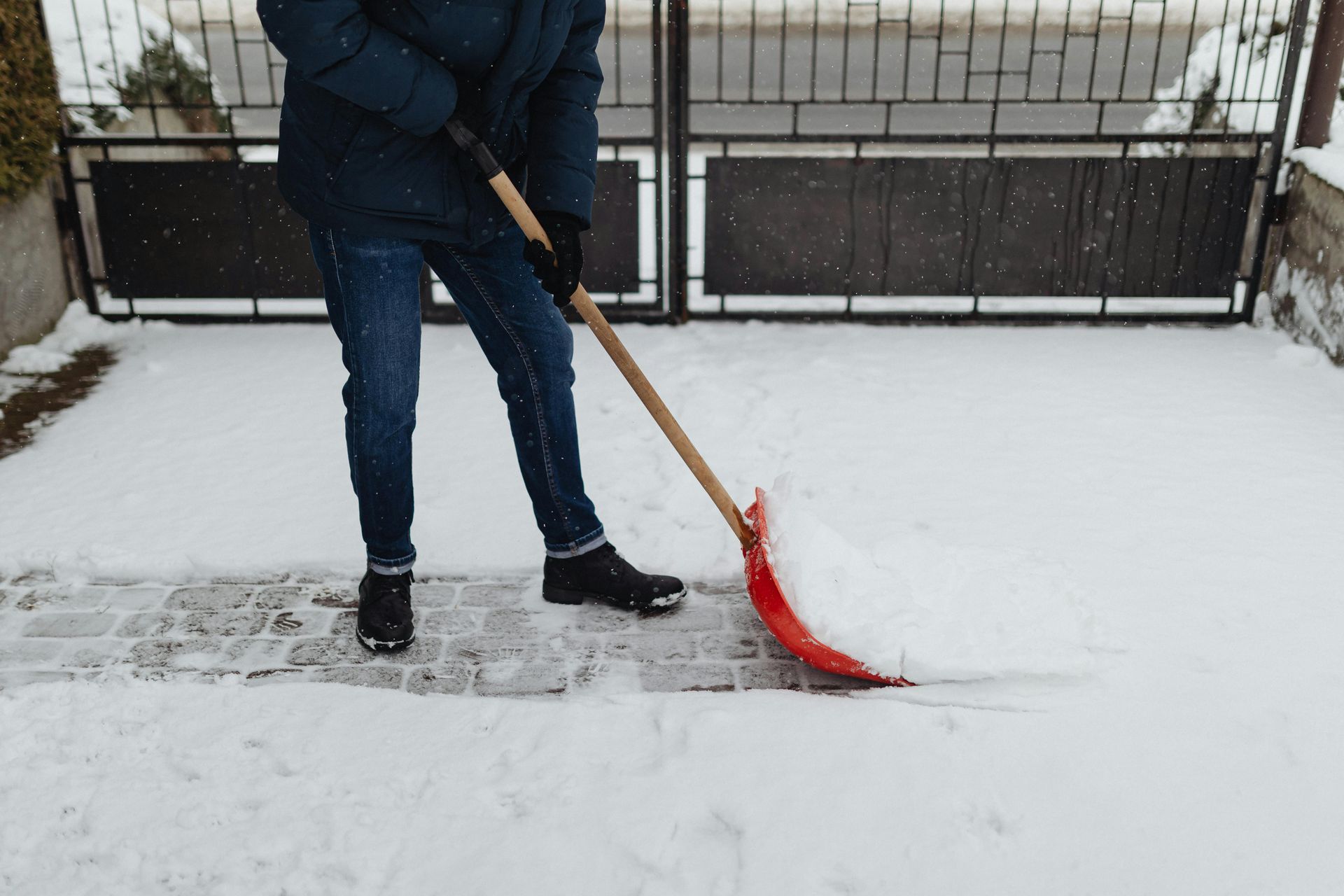 A person wearing a dark jacket and blue jeans uses a red-bladed shovel to clear snow from a paved walkway.