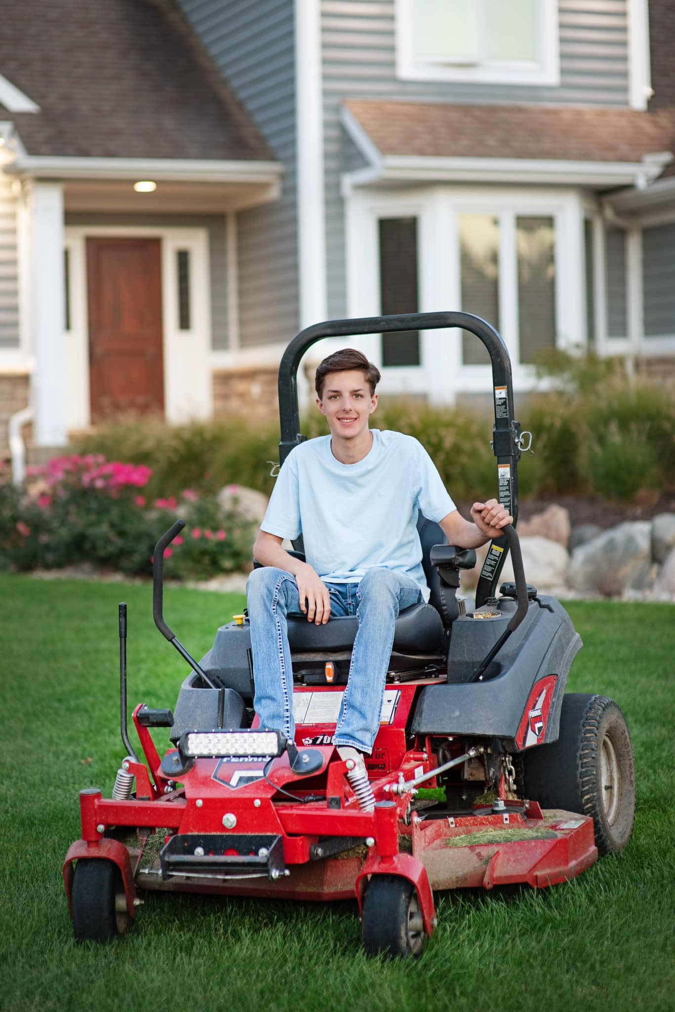 A young man is sitting on a lawn mower in front of a house.