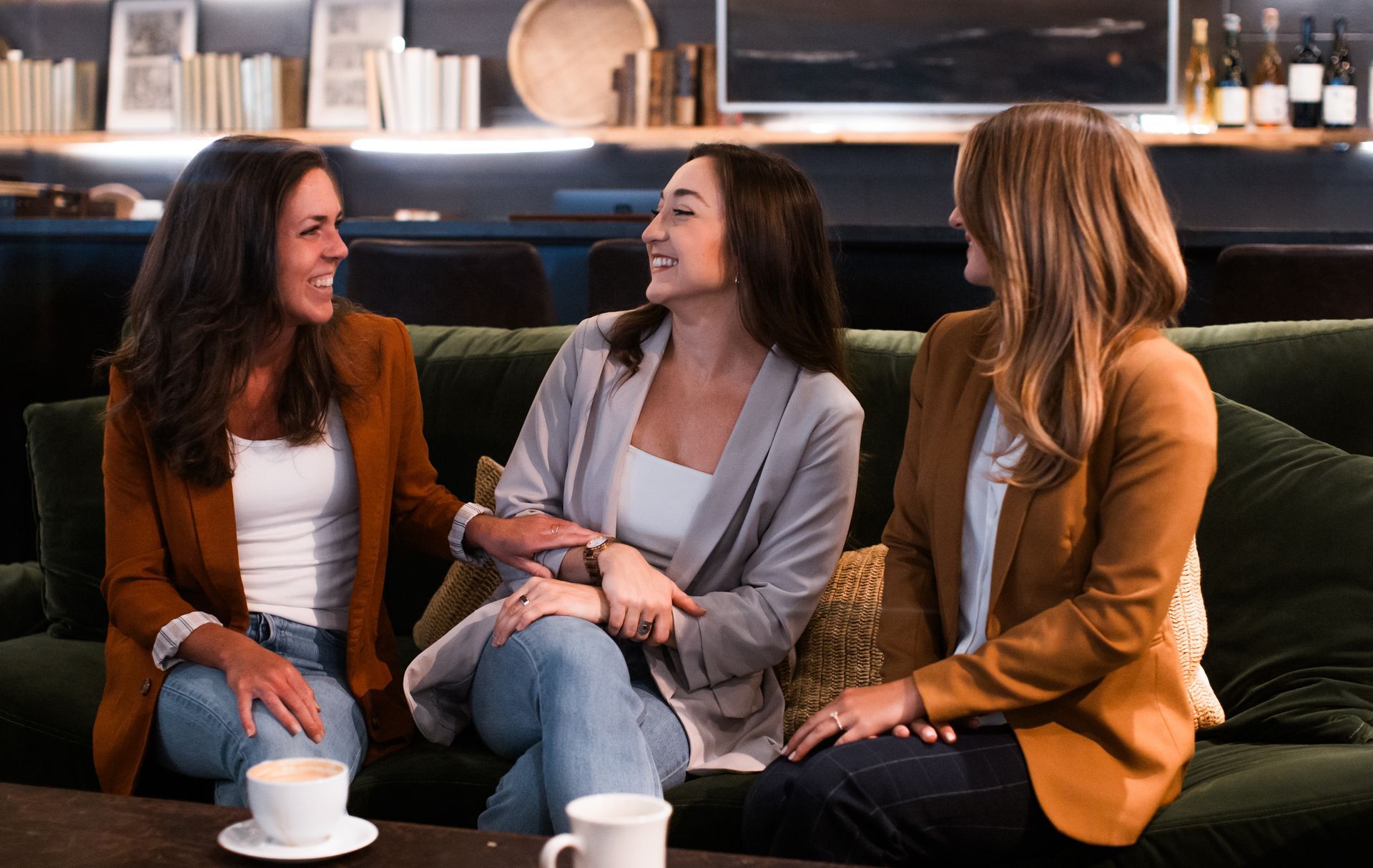 Three women are sitting on a couch in business attire and laughing