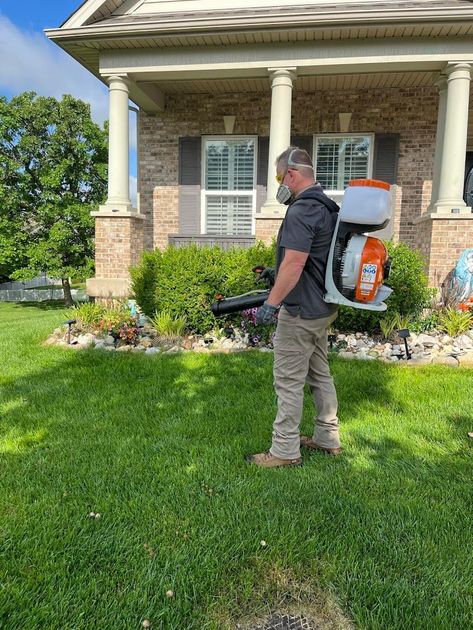 A man is spraying a lawn with a backpack sprayer in front of a house.