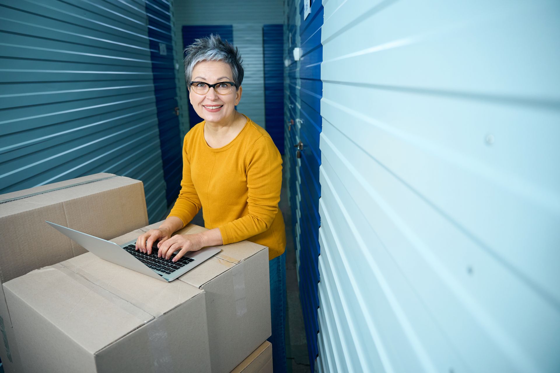 Woman using a laptop beside moving boxes inside a storage facility.
