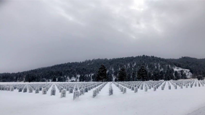 Snowy cemetery with rows of white headstones, mountain in the background under a cloudy sky.