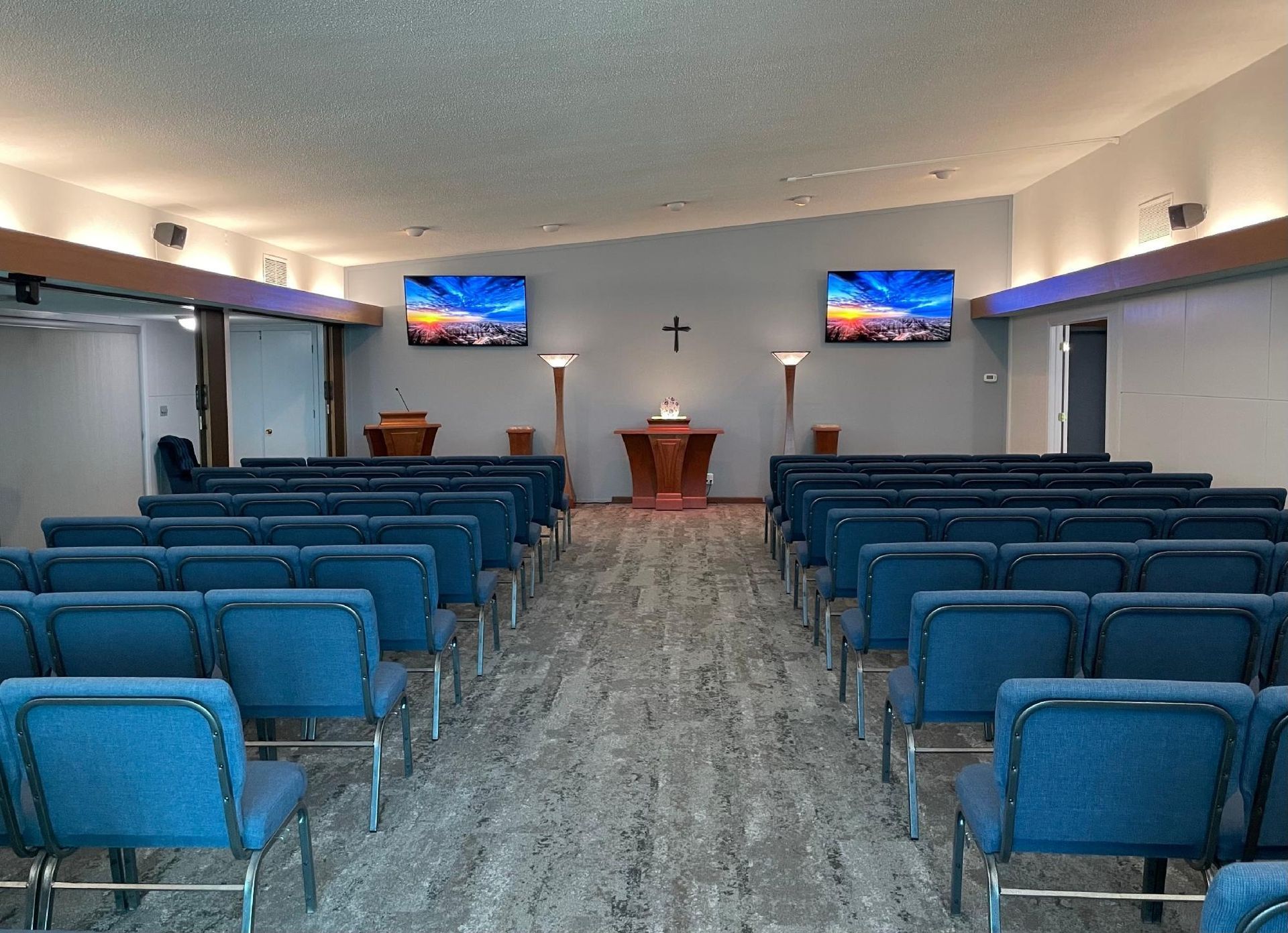 Interior of a chapel with blue chairs, a cross, and two screens displaying scenic views.
