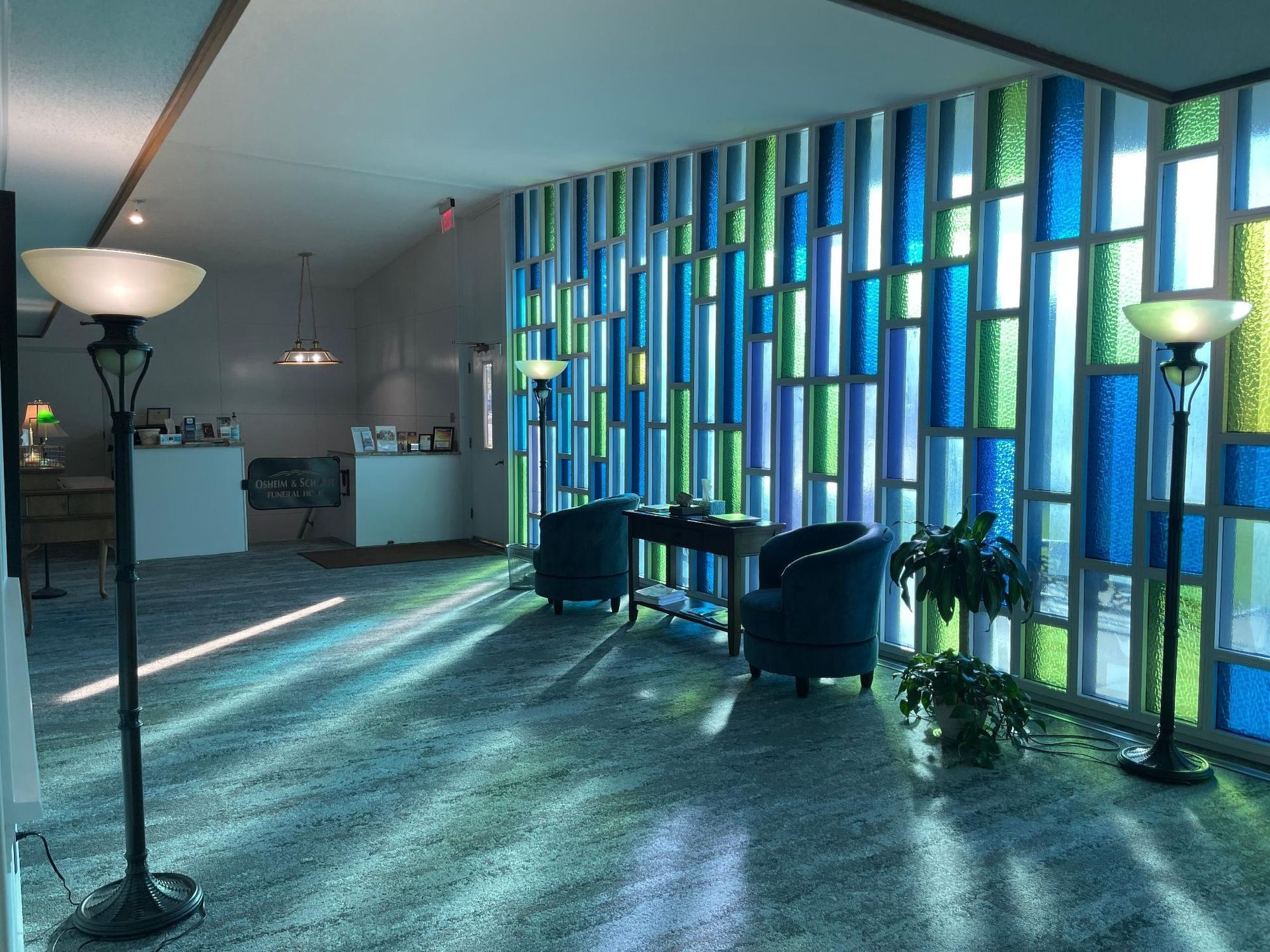 Lobby with blue, green, and white stained-glass wall, two armchairs, and floor lamps casting light.