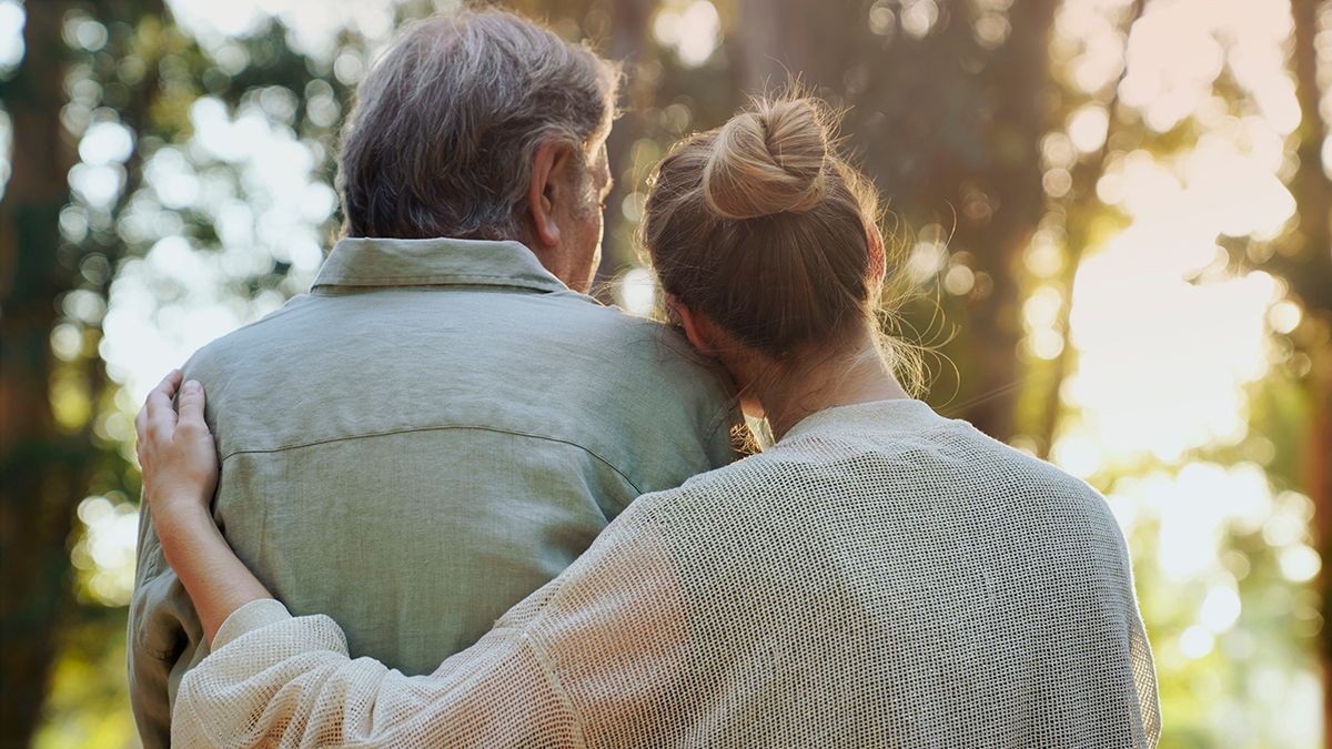 A person with their arm around another person's shoulders in a forest, sunlight filtering through the trees.