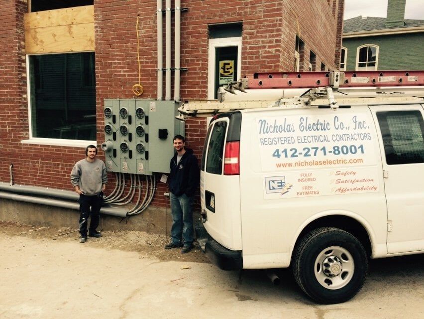 An electrical van by a brick building with workers next to outdoor meter panels An electrical van by a brick building with workers next to outdoor meter panels