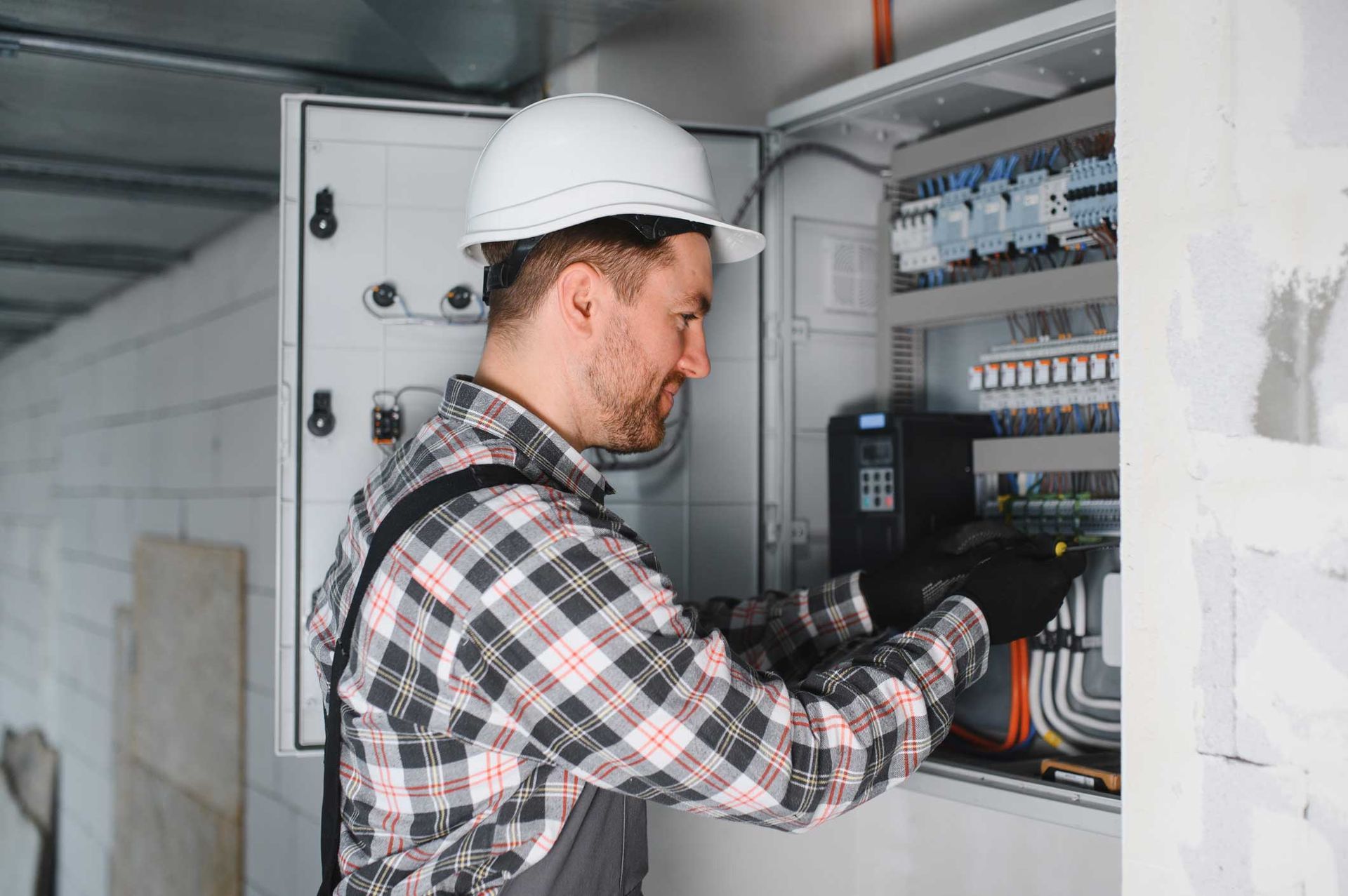 Electrician inspecting wiring in an open electrical panel, wearing a hard hat and gloves.