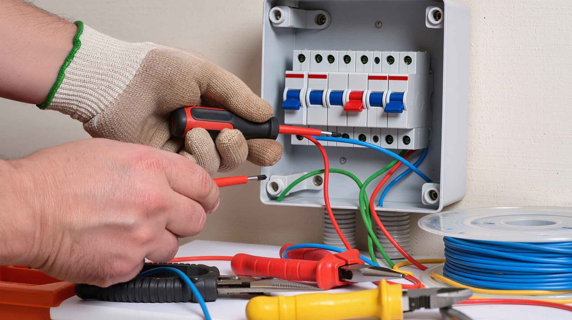 Gloved hands wiring a small electrical panel with circuit breakers and colorful cables