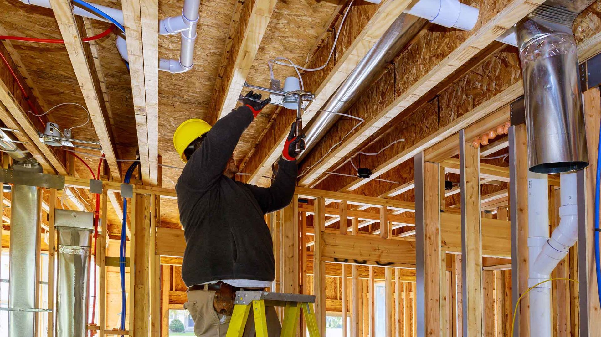 Worker fastening pipes in a framed house under construction, wearing a hard hat and safety vest