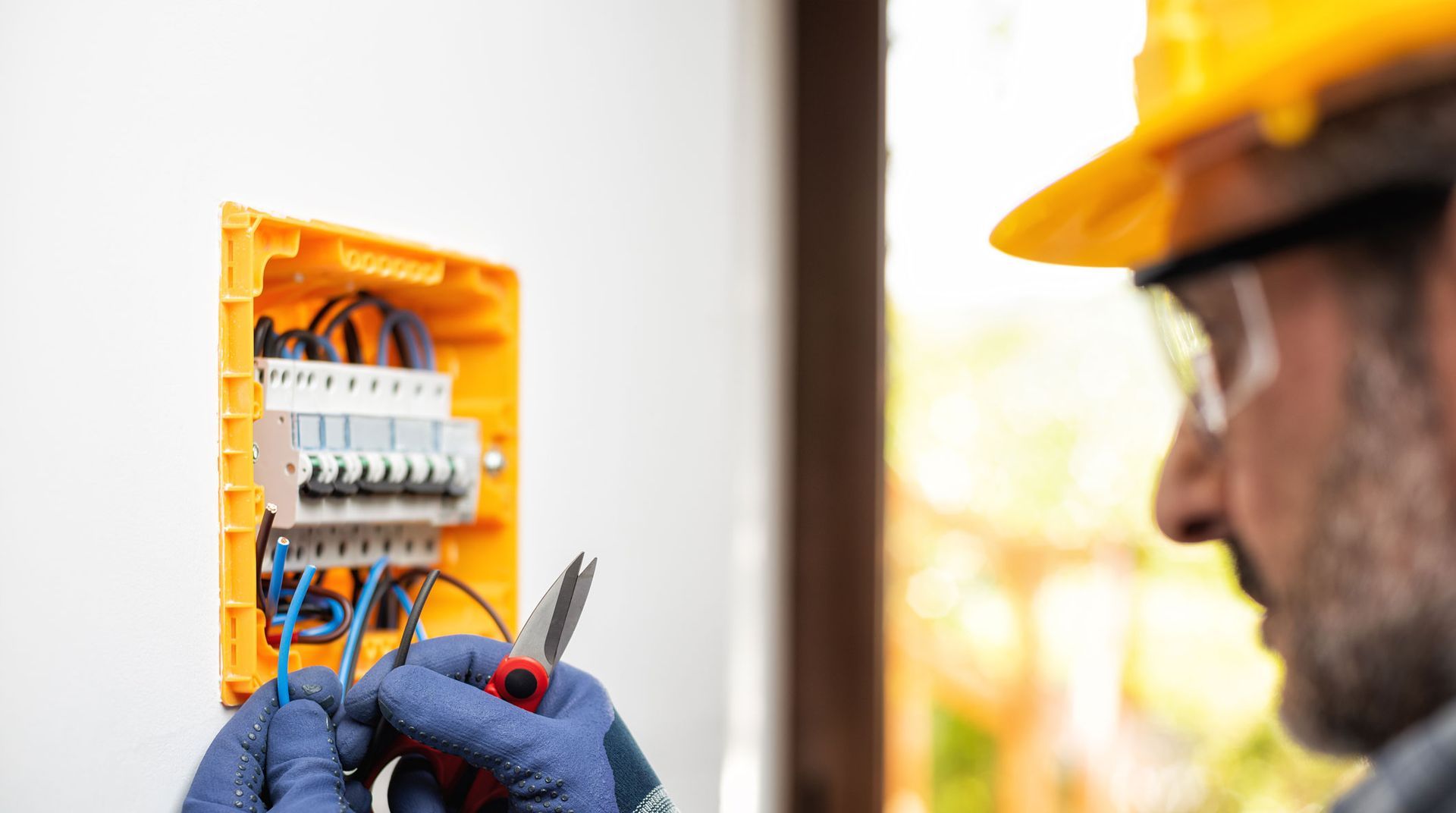 Electrician testing wiring in an orange electrical panel with a multimeter, wearing gloves and a hard hat.
