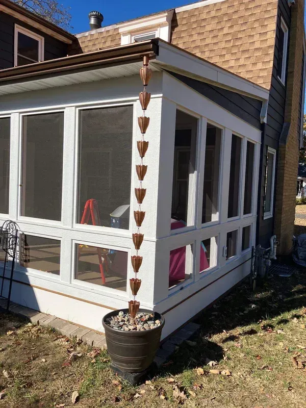 A copper rain chain hangs from the gutter of a white screened-in porch, emptying into a large pot filled with rocks.