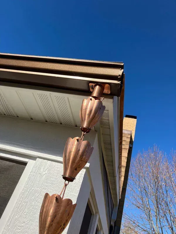 A copper rain chain hangs from the corner of a brown gutter on a white house.
