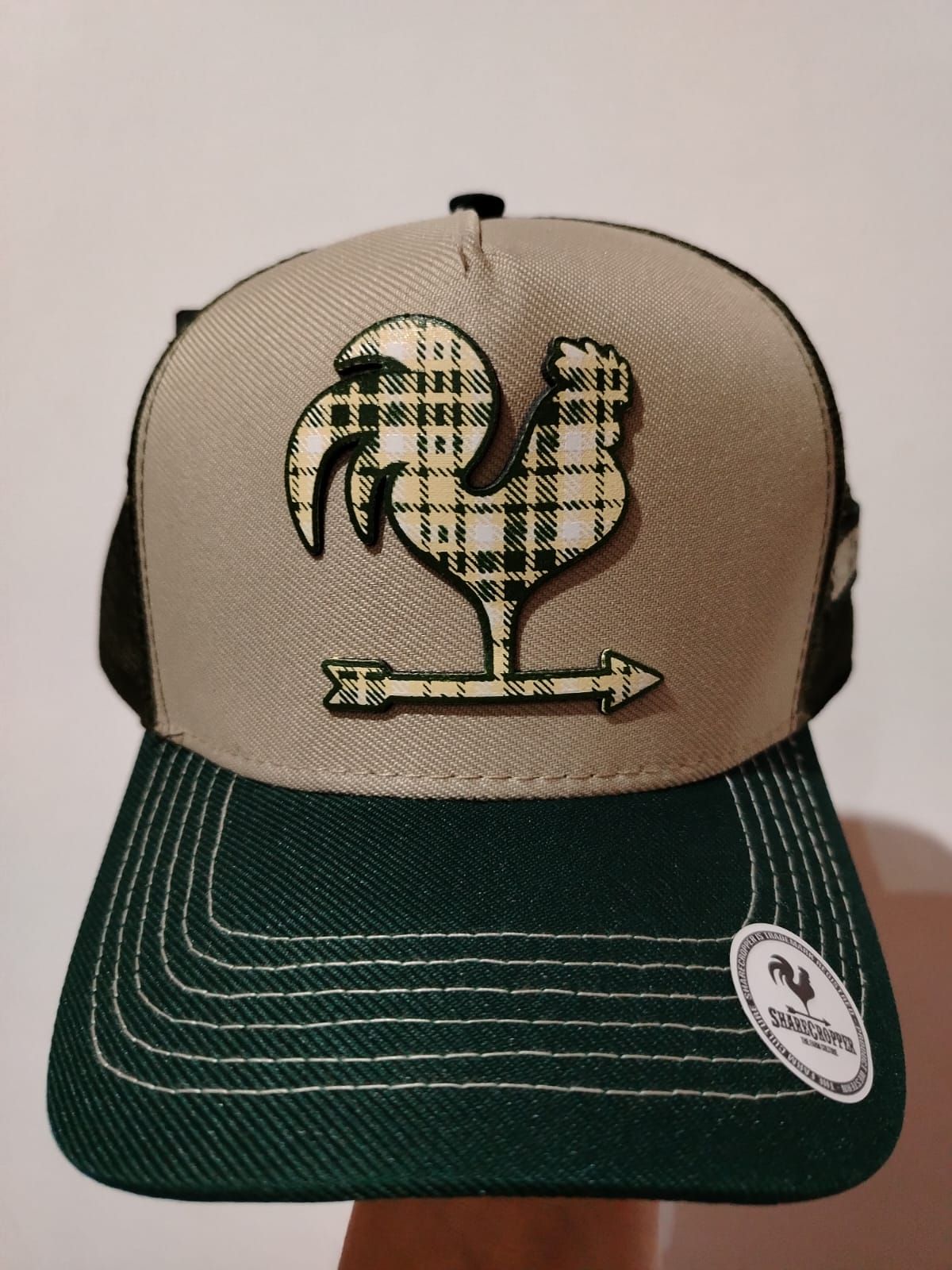Gorra de camionero verde y tostado con un emblema de gallo a cuadros y una flecha.