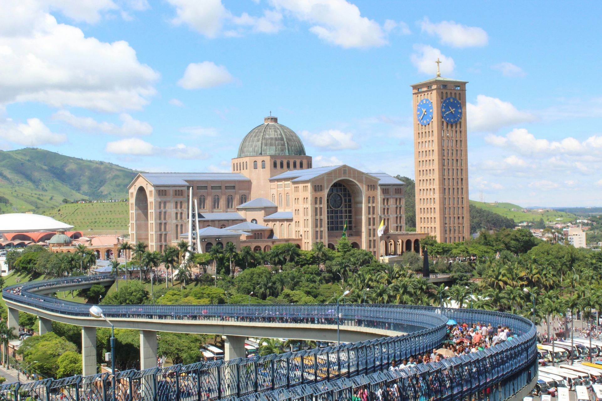 Basílica de Aparecida, Brasil, com pessoas em uma passarela. Grande edifício bege com torre do relógio e uma cúpula verde.